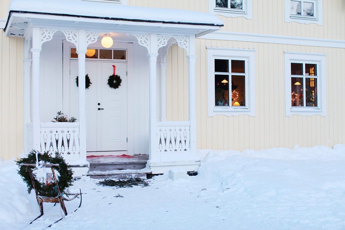 Verschneiter Vorgarten mit weihnachtlich dekoriertem Schlitten, vor hellgelbem Schwedenhaus mit überdachtem Hauseingang