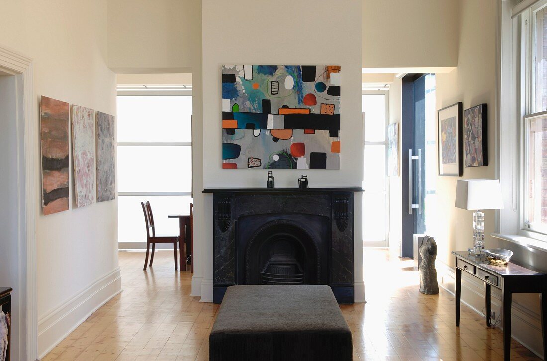 Modern artwork above central open fireplace flanked by open doorways with further artworks and console table; ottoman in foreground