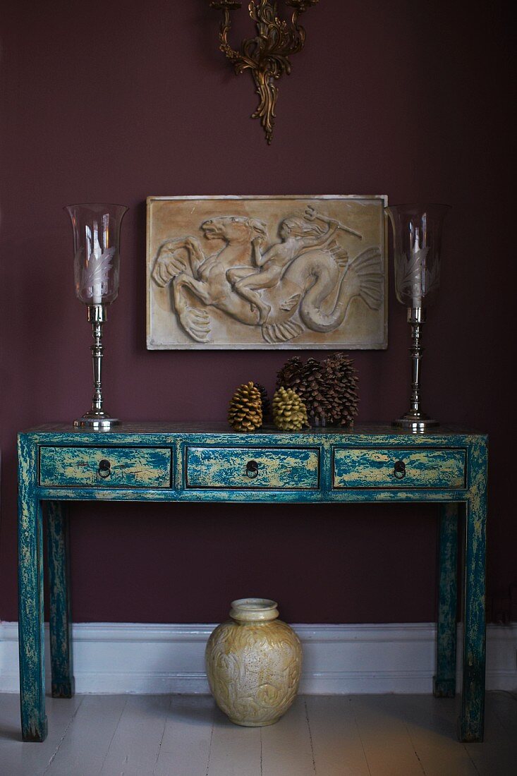 Elegant, silver candlesticks on vintage-style console table with drawers below antique low-relief on aubergine-coloured wall
