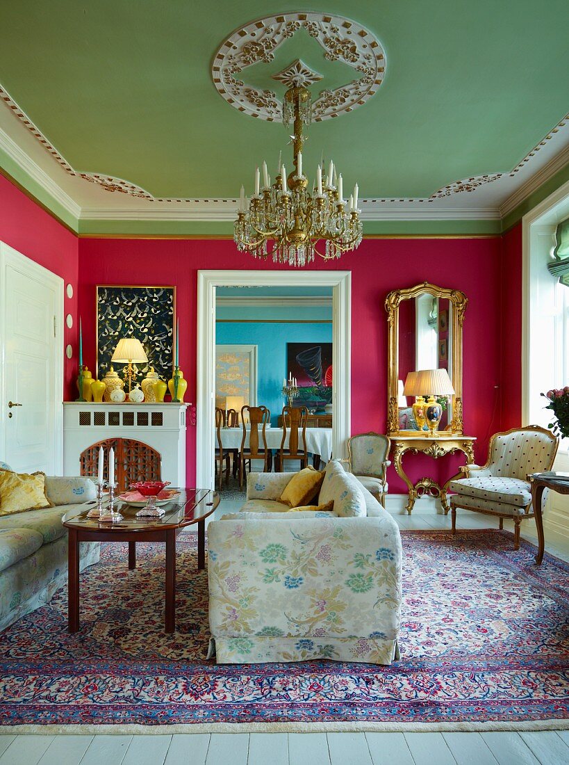 Grand salon with deep pink walls, pastel green stucco ceiling, chandelier above seating area and view of dining table through open doorway in background