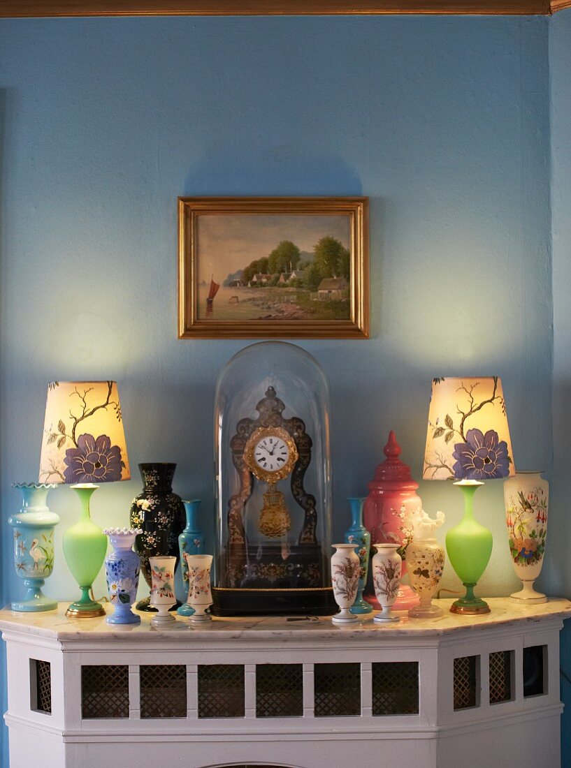 Verschiedene bemalte Vasen und Tischuhr mit Glashaube auf verkleidetem Kamin vor hellblau getönter Wand mit Landschaftsbild in Goldrahmen