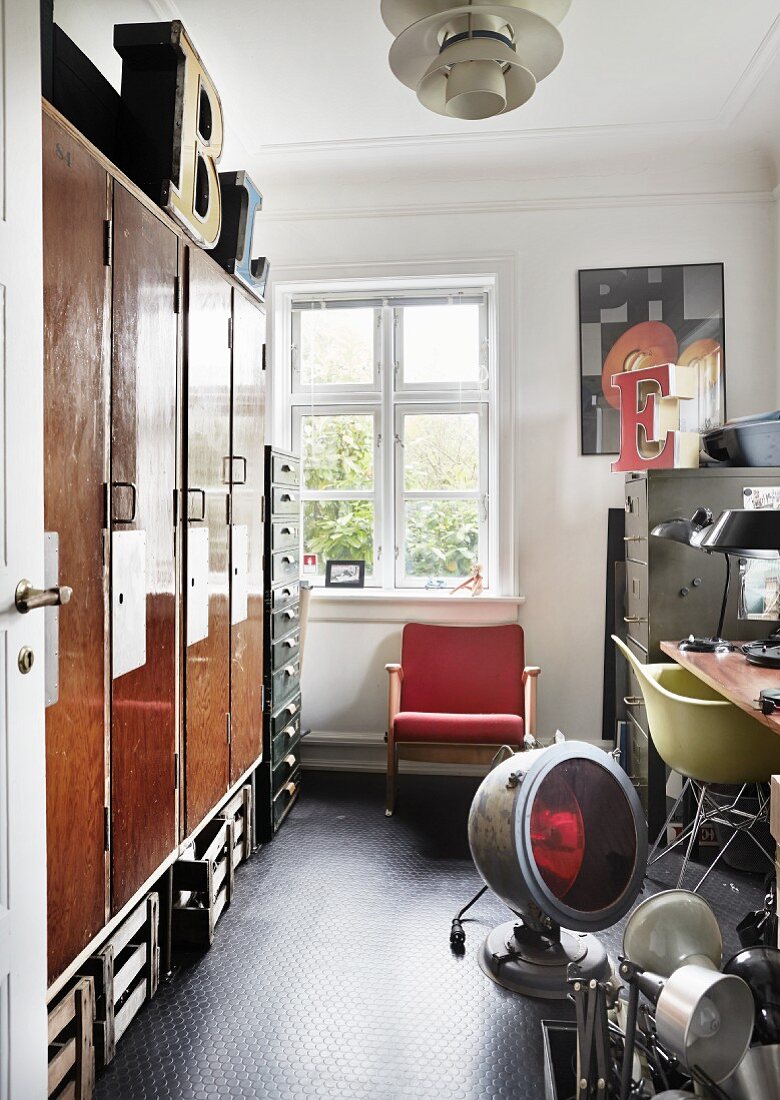 Open door with view into workshop; vintage lamps on floor, old fitted cupboards and retro armchair below window