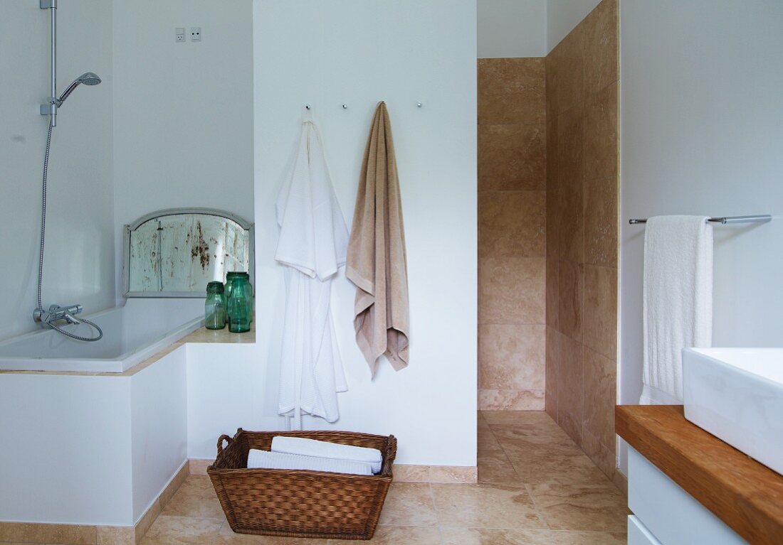 Wäschekorb auf Boden zwischen eingebauter Badewanne in Nische und Duschbereich mit beigefarben, marmorierten Fliesen auf Wand und Boden