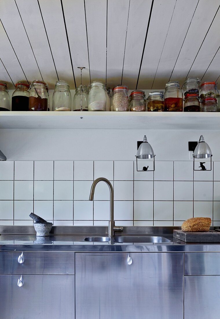 Ausschnitt einer Küchenzeile mit Spüle aus Edelstahl, vor weissen Fliesen an Wand, darüber Ablage mit Vorratsgläsern