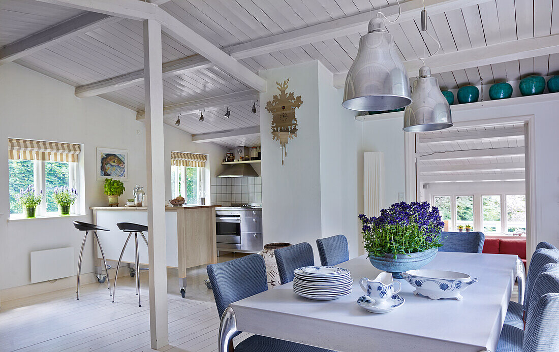 Crockery and bowl of flowers on dining table below pendant lamps with retro metal lampshades hanging from white wooden ceiling; open-plan kitchen with bar stools at counter