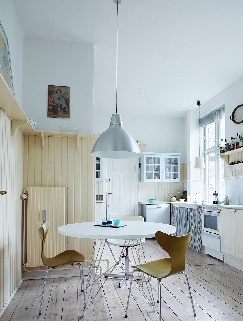 Dining area with classic-style shell chairs below retro, metal pendant lamp in rustic kitchen with cream wood panelling