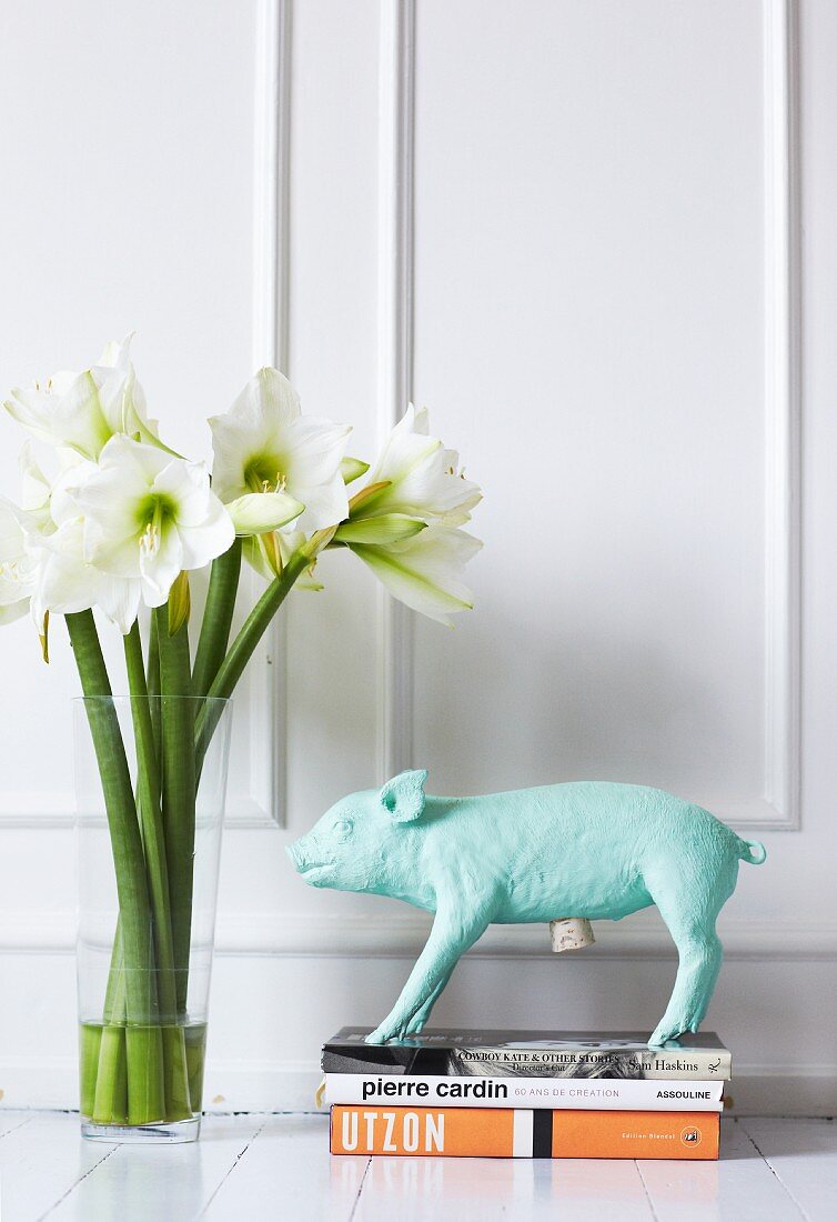 Pig figurine on stacked books and glass vase of flowers against of white, wood-panelled wall