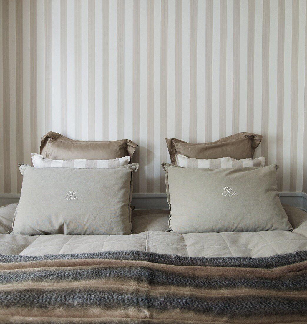Kissenstapel in abgestimmten Naturtönen auf französischem Bett, vor Wand mit Streifentapete in traditionellem Schlafzimmer mit dezenten Farbtönen
