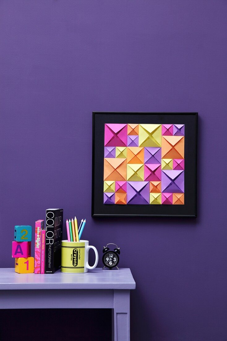 DIY-Bild mit 3D-Effekt aus Origami-Quadraten verschiedener Größen und Farben vor blauvioletter Wand; Schreibtisch mit farblich abgestimmter Deko