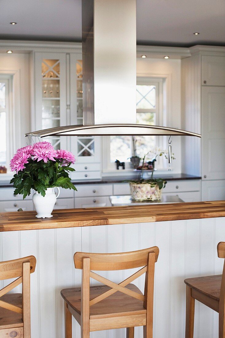 Wooden bar stools at kitchen counter below stainless steel extractor hood in open-plan, country-house kitchen