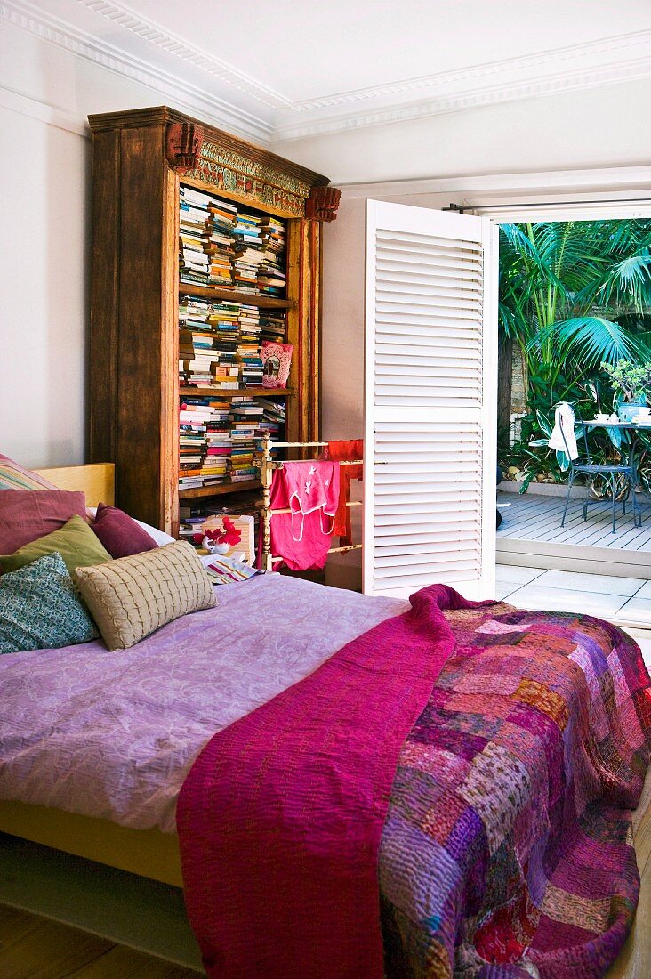 Patchwork-Plaid auf Doppelbett, neben antikem Bücherschrank, gegenüber offene Terrassentür mit Blick auf Palme