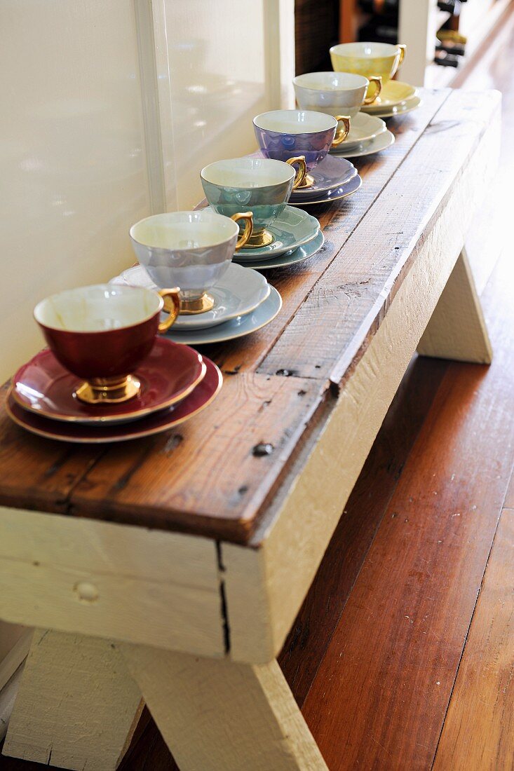 Espresso cups of various colours with gilt handles lined up on rustic wooden bench