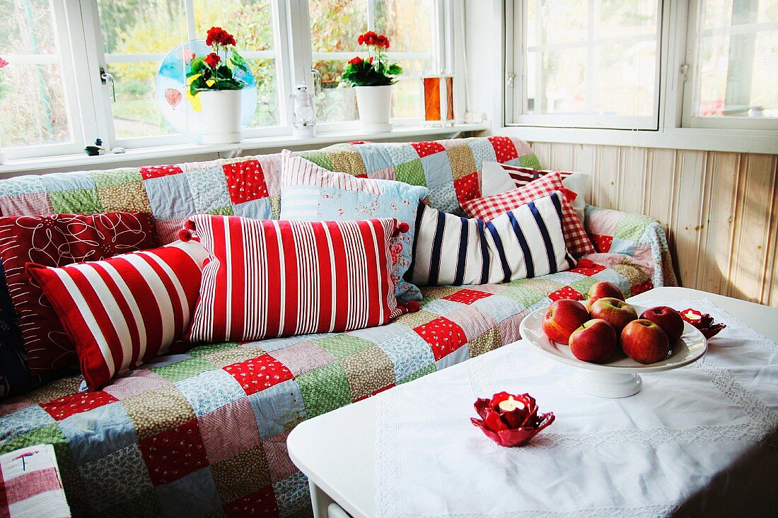 Gemütliche Ecke in der Loggia - Schale mit Äpfeln auf weißem Tisch, vor Sofa mit Patchworkdecke und gemusterten Kissen