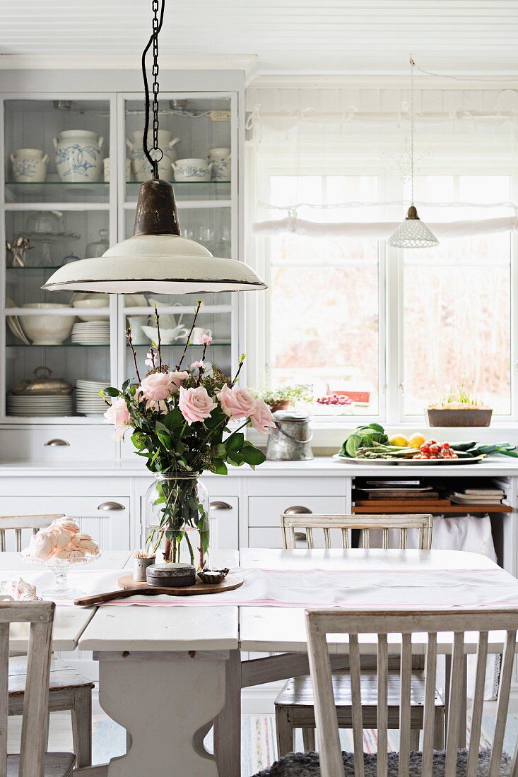 Nostalgic kitchen lamps above glass vase of roses on dining table in white, Swedish country-house kitchen