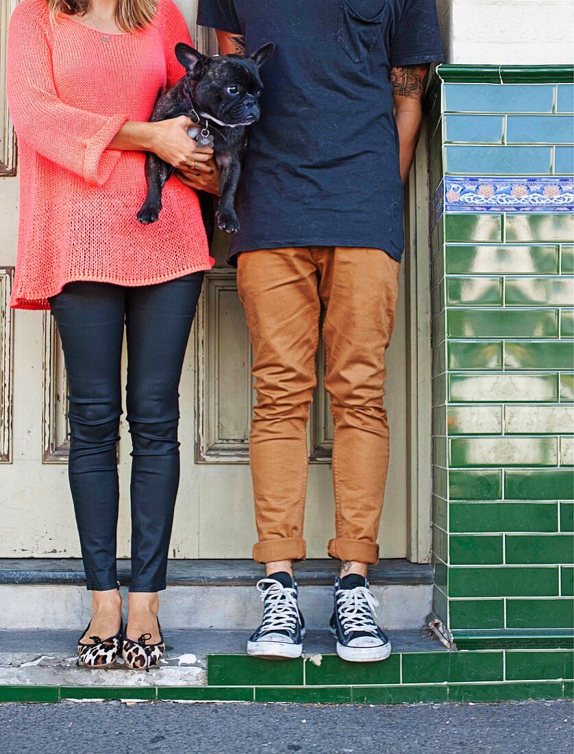Beine und Körper eines jungen Paares, mit Hund im Arm, vor Haustür