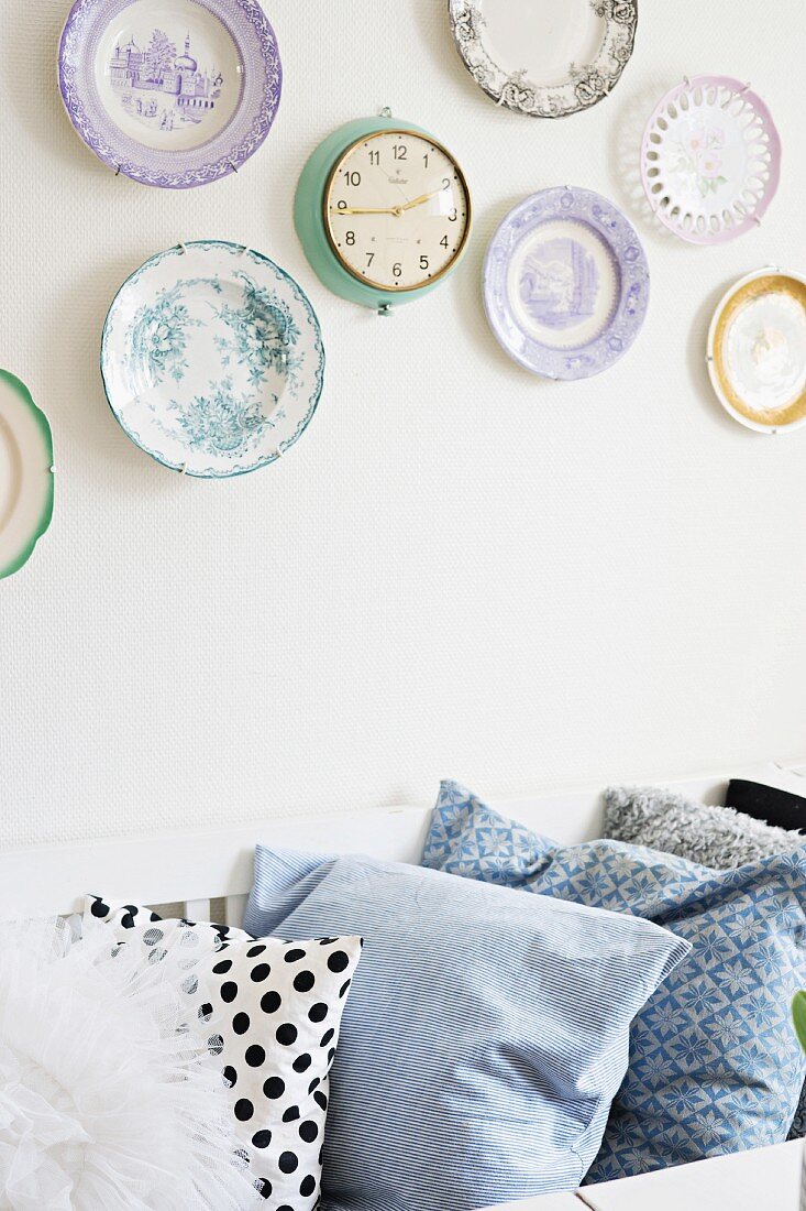 Row of plain and patterned scatter cushions below collection of plates and retro clock on wall