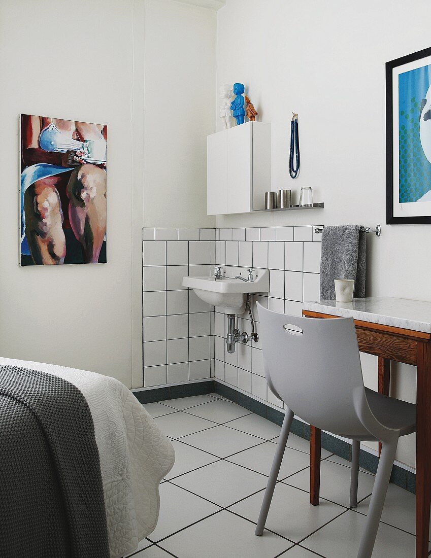 Grauer Kunststoffstuhl vor Tisch, neben minimalistischem Waschbereich in Schlafzimmerecke mit modernem Bild an Wand