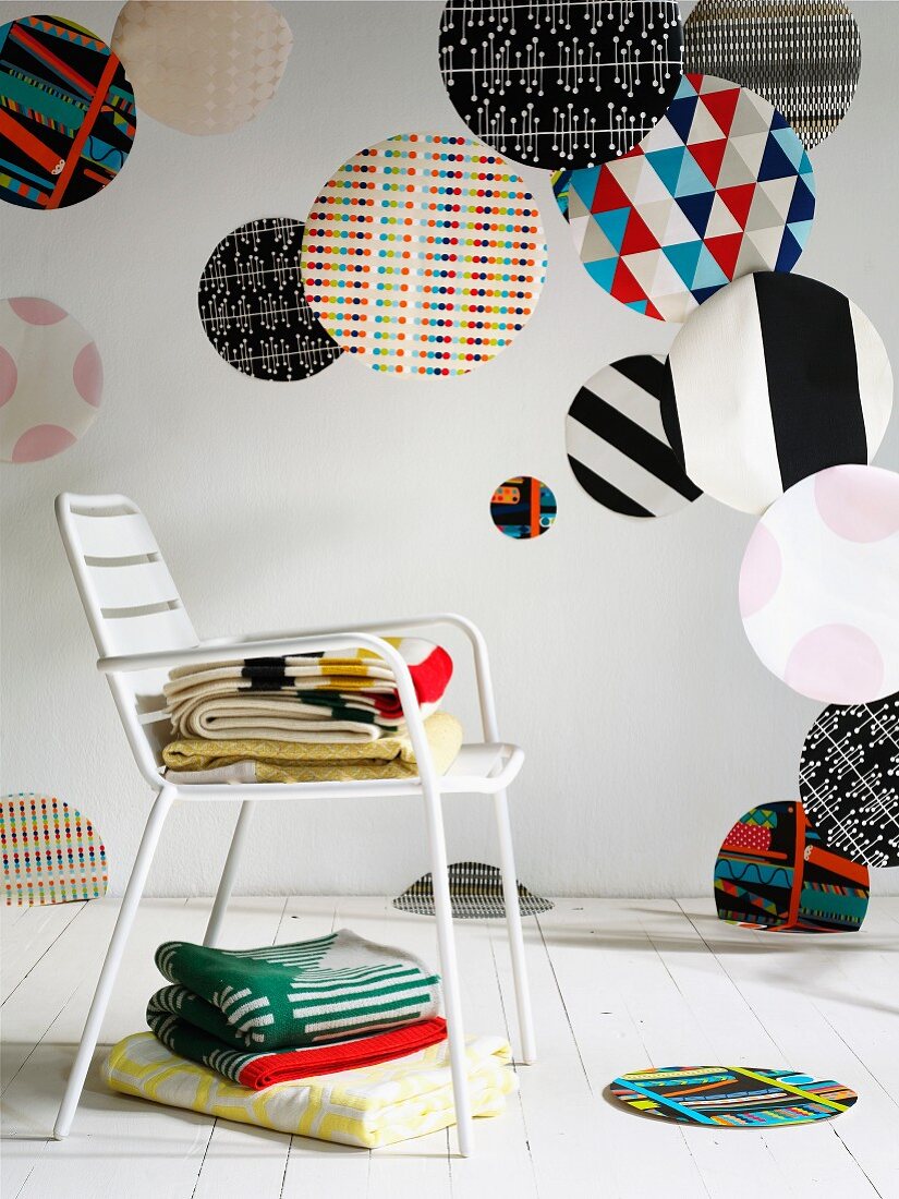 Various large colourful fabric circles of various patterns hanging in interior, stacked blankets on white metal chair and white wooden floor