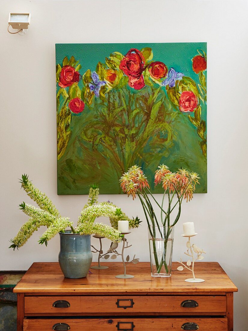 Blumen in Vasen und Kerzenständer mit weissen Stumpenkerzen auf Holzkommode vor Wand mit gemaltem Blumenbild