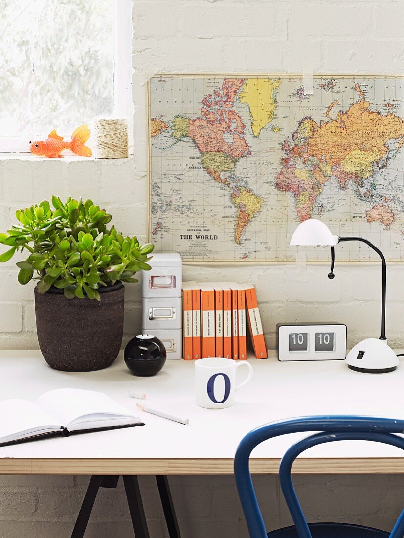 Money tree in dark pot and office utensils next to white desk lamp on table below map of world hung on whitewashed brick wall