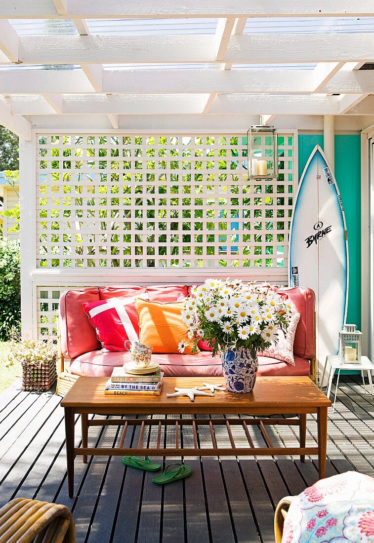 Margeritenstrauss auf Couchtisch vor gemütlicher Bank mit Kissen auf der Veranda eines Strandhauses, dahinter weiße Holz Sichtschutzwand