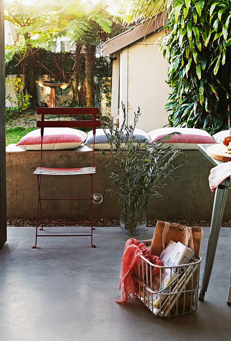 Einkaufskorb auf Terrassenboden und schlichter Metall Klappstuhl vor Brüstungsmauer mit gestreiften Kissen, dahinter Palmen im Innenhof