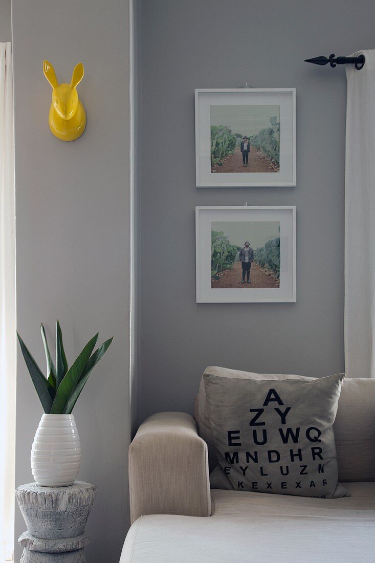 Sehtest-Tafel als Deko-Print auf einem Sofakissen; weiß gerahmte Bilder und gelbe Deko-Hasentrophäe an hellgrau getönter Wand