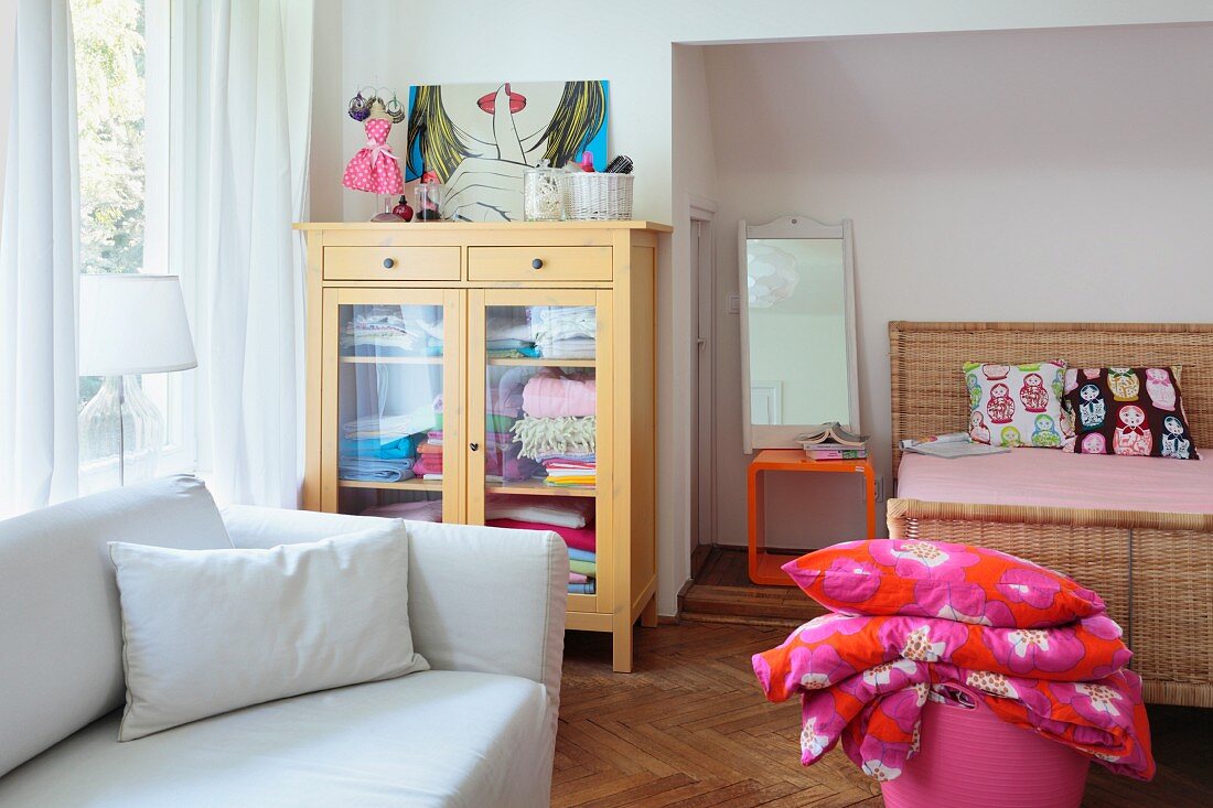 Schlafzimmer mit weißem Sofa und nostalgischem Wäscheschrank mit Glastüren vor Fensterseite, im Hintergrund Doppelbett aus Rattan in Nische und pink-orangefarbener Bettwäsche in Korbtasche