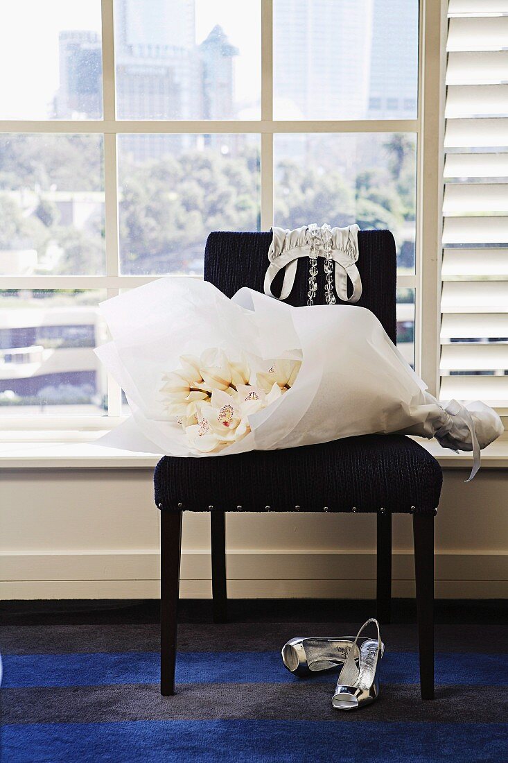 Bouquet of white flowers wrapped in paper on black upholstered chair and sling-back high heels on carpet below lattice window