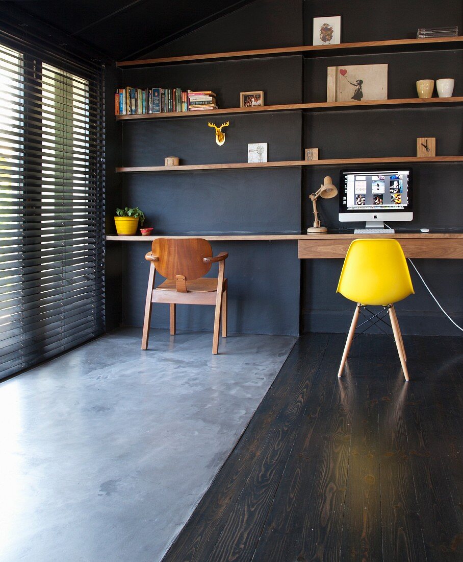 Gelber Klassiker Schalenstuhl vor minimalistischem Schreibtisch unter Regalablagen aus Holz an schwarz getönter Wand, Boden im Materialmix, Estrich- und Holzbelag