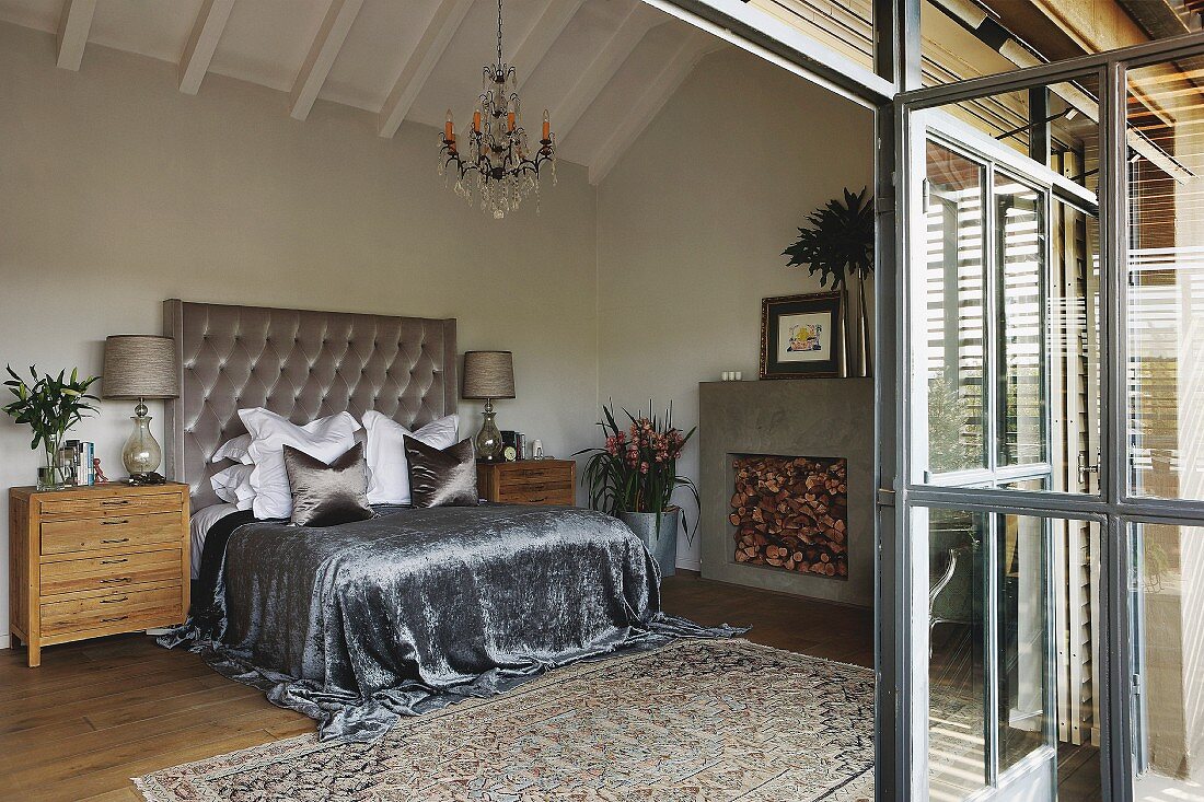 Spacious attic bedroom; shimmering silver throw on double bed with tall, button-tufted headboard against wall