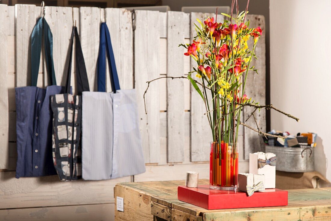 Freesien in mit Strohhalmen dekorierter Glasvase, im Hintergrund Stofftaschen an Bretterwand