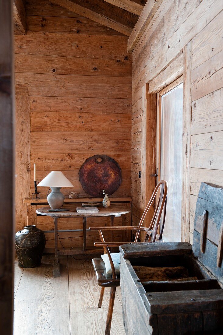 Offene Holztruhe neben Stuhl, im Hintergrund schlichter Tisch mit Tischlampe im Wohnzimmer einer Hütte
