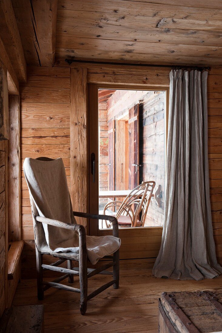 Rustikaler Armlehnstuhl mit Decke neben verglaster Terrassentür in Zimmerecke einer Holzhütte