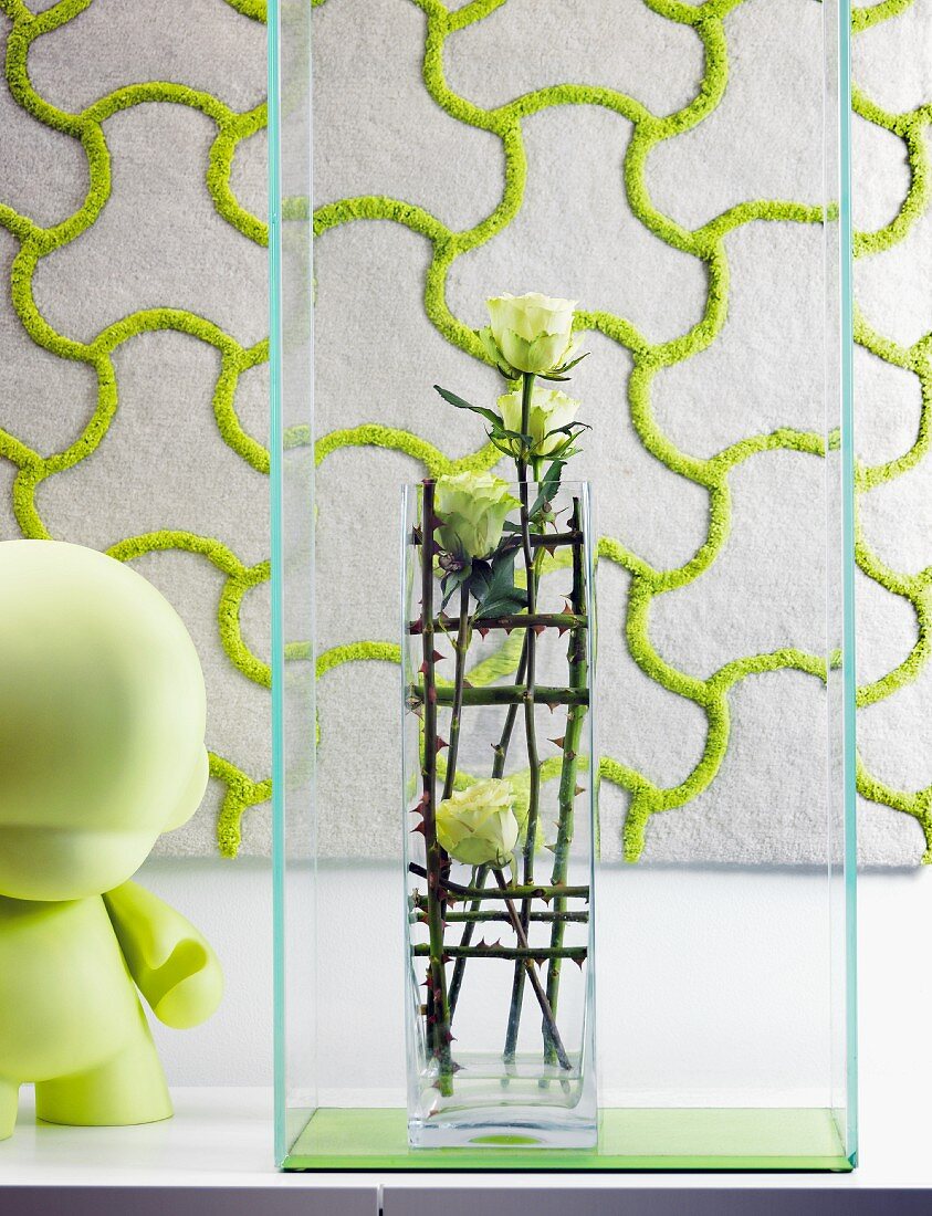 Glasvase mit Rosengesteck in Glasbox neben grüner Spielzeugfiur; textiles, gemustertes Wandpaneel im Hintergrund