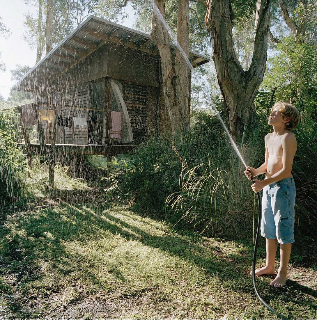 Junge mit Wasserschlauch beim Giessen im Garten, im Hintergrund Waldhütte mit Pultdach