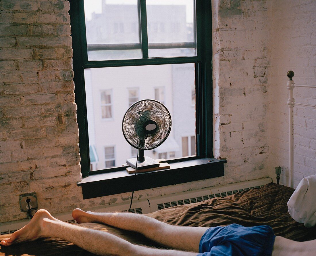 Mann im Bett liegend, vor rotierendem Ventilator auf Fensterbank