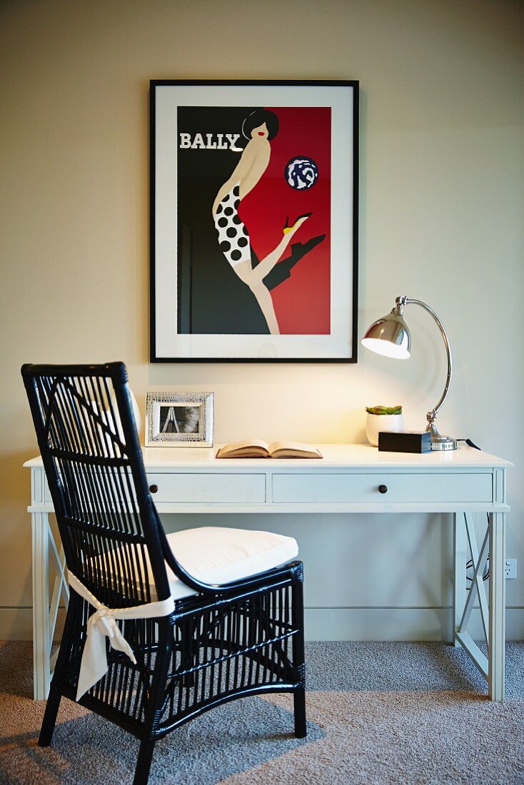 Schwarz lackierter Rattanstuhl vor weißem Schreibtisch mit Lampe und Poster darüber an der Wand