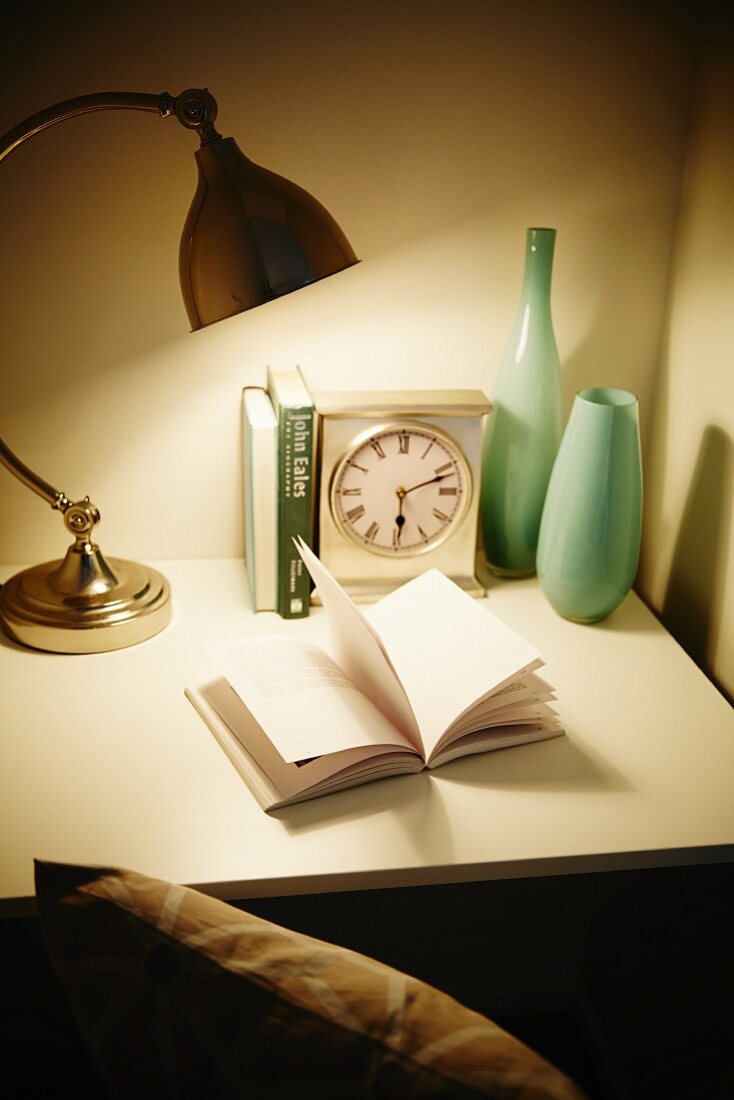 Aufgeschlagenes Buch unter einer Lampe auf Schreibtisch mit Uhr und Vasen