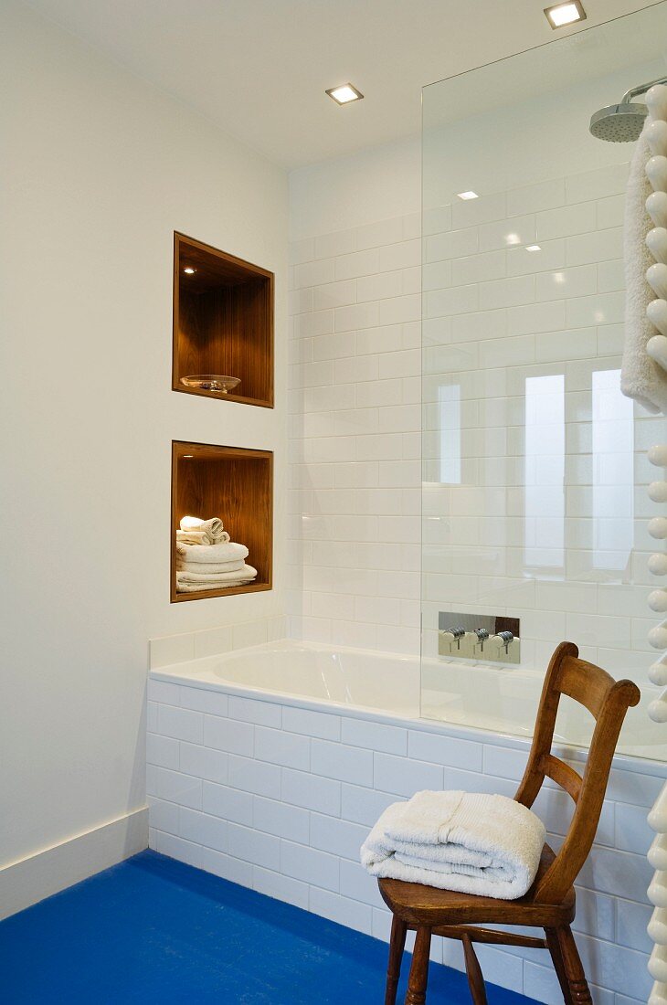 Minimalist, white, designer bathroom with towels in niches, glass shower screen, blue floor and rustic wooden chair
