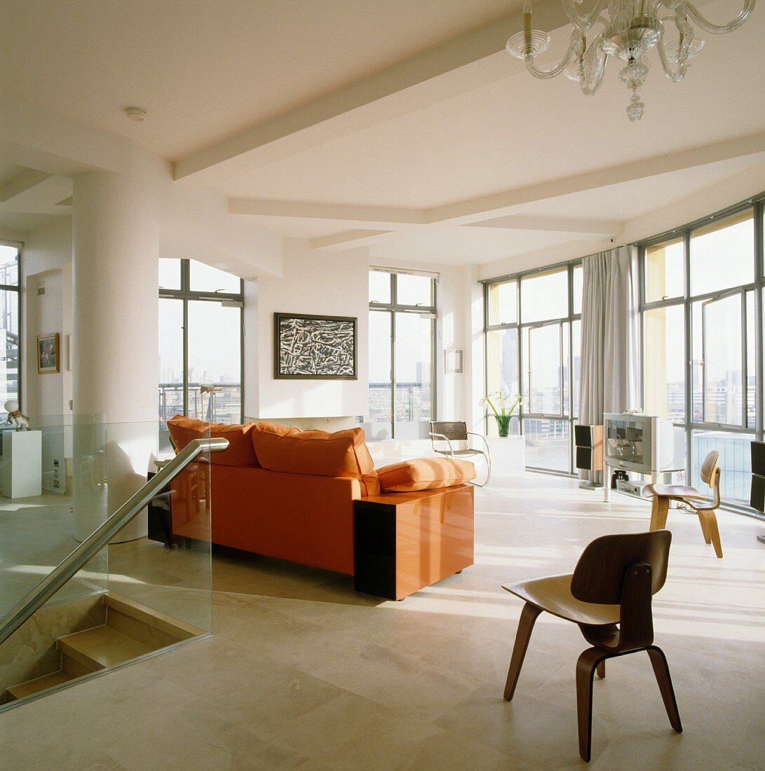 Wooden designer chair and sofa with orange upholstery in living room of penthouse apartment with floor-to-ceiling windows
