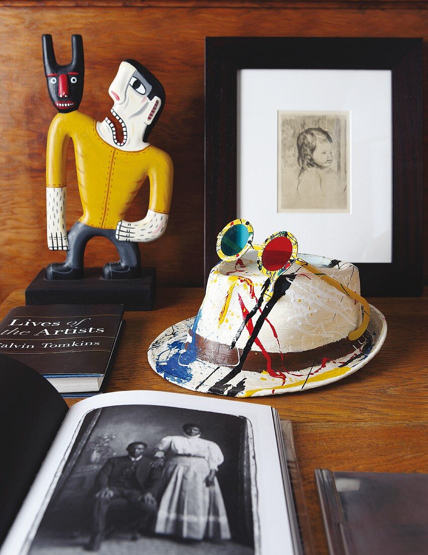 Bemalter Hut mit aufgesetzter Sonnenbrille, Bücher und bemalte Holzfigur auf Tisch
