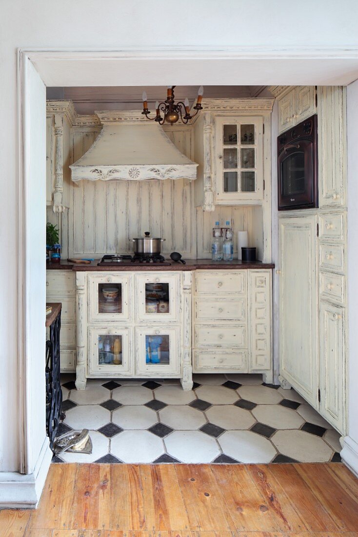 Open doorway with view into shabby-chic kitchen area with white cupboards, some with lattice doors, and white-tiled floor with black accent tiles