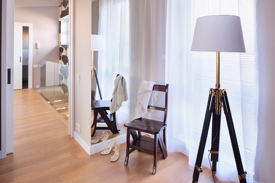 Standard lamp with retro, wooden tripod base and white lampshade and vintage wooden chair in front of window with floor-length, translucent curtain