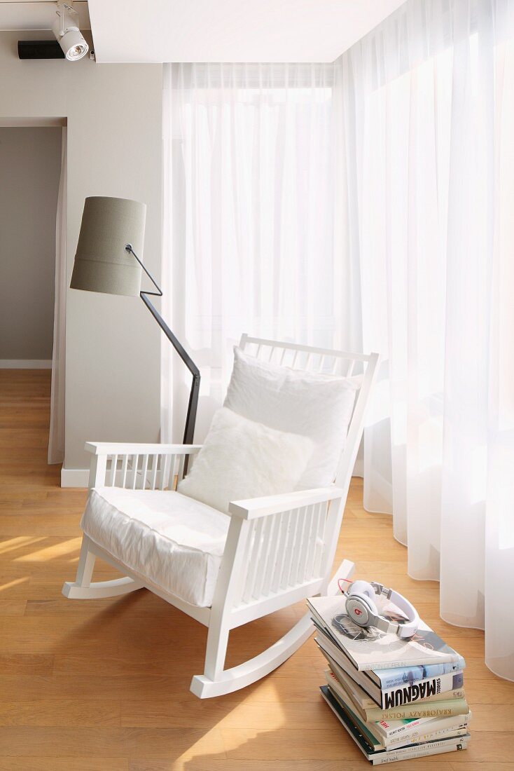 White, wooden rocking chair with white cushions and standard lamp with grey lampshade in front of floor-length, translucent curtains on windows