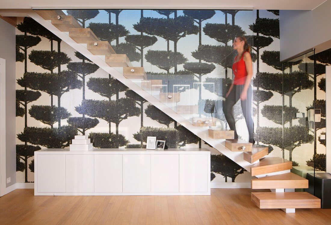 Frau auf Treppenaufgang mit Massivholzstufen und Glasbrüstung, darunter weisses Sideboard an tapezierter Wand mit Baummotiven