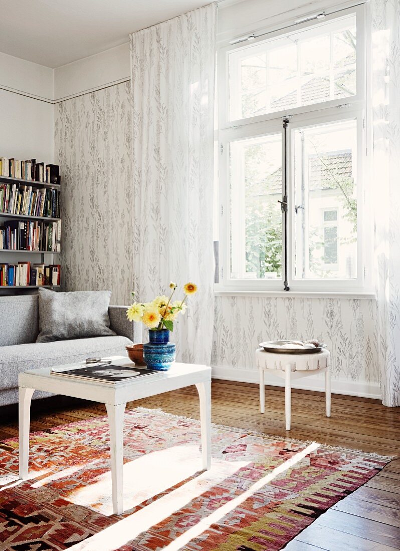 Vase of flowers on white side table on rug in front of window with floor-length, translucent curtains; sofa and bookshelves against wall in background