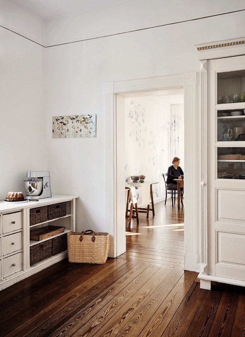 Traditional country-house kitchen with white-painted cupboards and woman sitting at dining table seen through open door