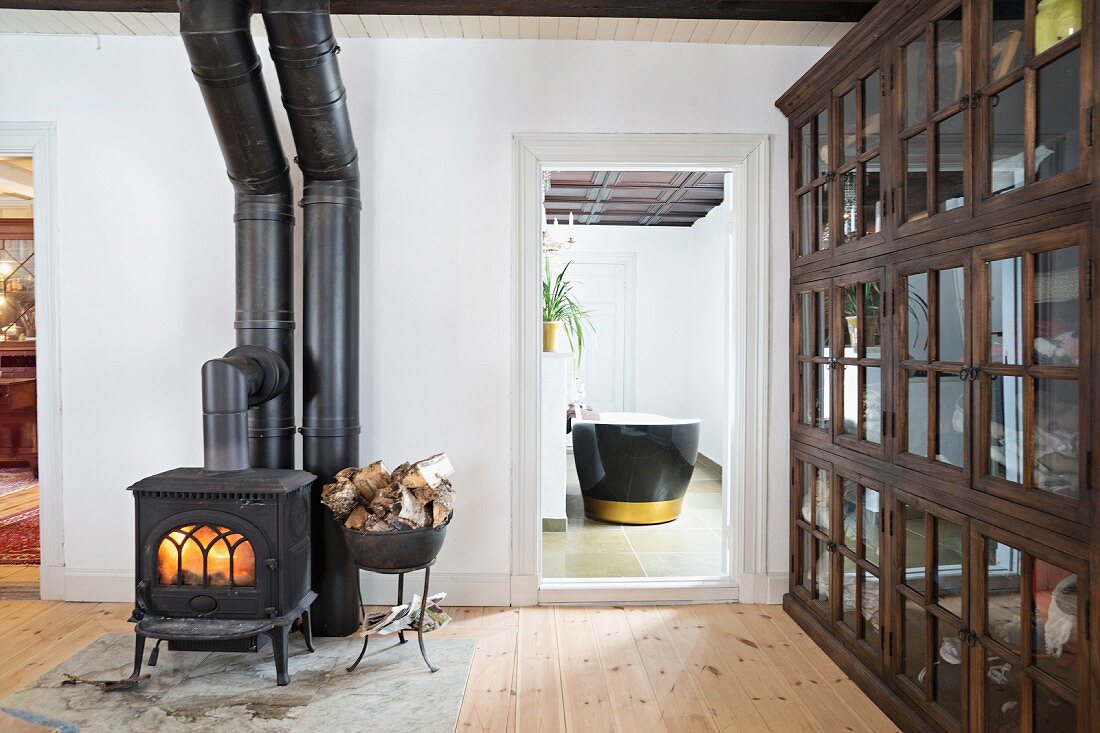 Schwarzer Kanonenofen und Holzlager, seitlich Vitrinenschrank mit Sprossentüren in schlichtem Wohnzimmer, offene Zimmertür und Blick auf Badewanne