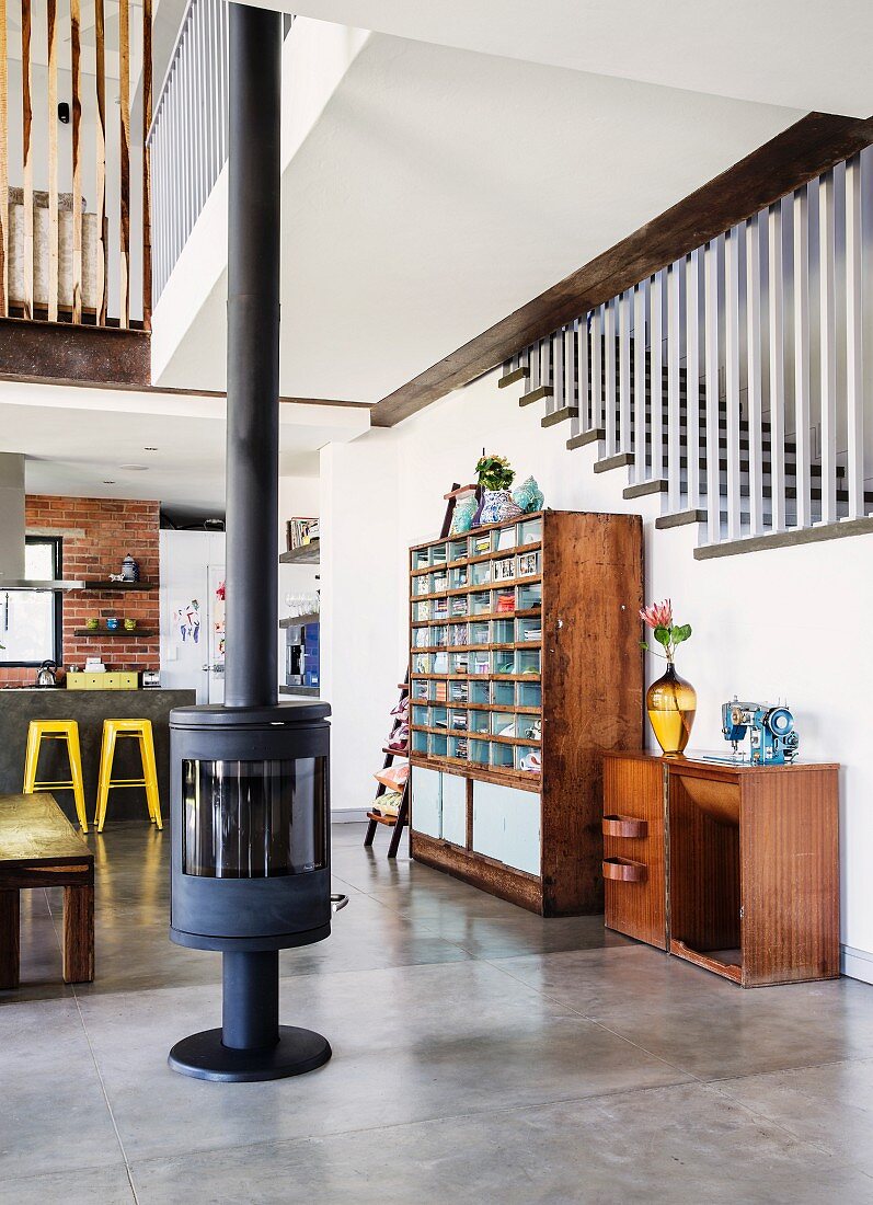 Freistehender Kaminofen auf Estrichboden, seitlich halbhoher Schrank und Vintage Regalschrank an Treppenhauswand in offenem Wohnraum